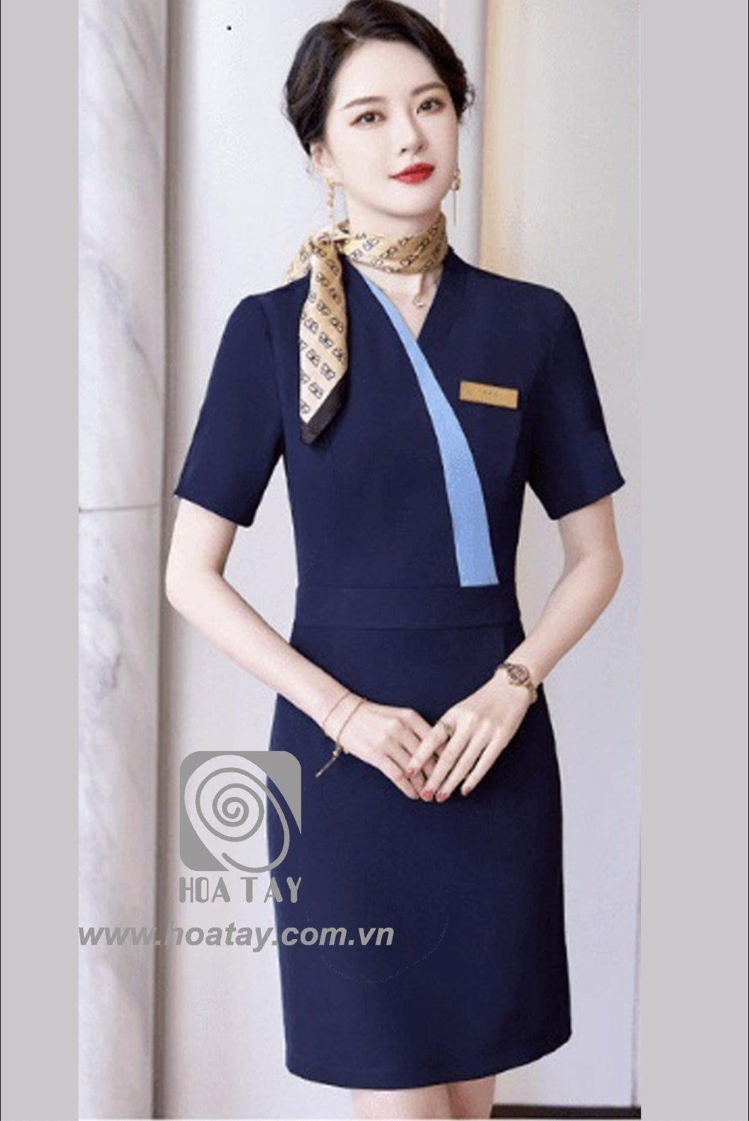 Một nữ nhân viên đứng thẳng, mặc váy đồng phục liền thân (dress) màu xanh navy, cổ chữ V phối viền xanh nhạt, tay ngắn, chất liệu vải đứng form, đeo khăn quàng cổ họa tiết, bối cảnh văn phòng ánh sáng