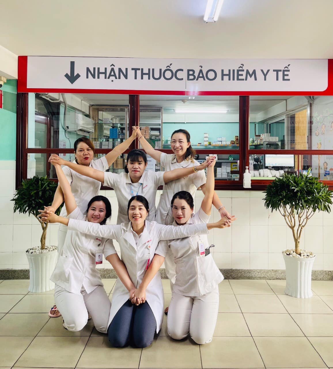 Sáu nữ nhân viên y tế (6 người) đang tạo dáng hình tháp, mặc áo blouse trắng (áo khoác bác sĩ) cổ bẻ, chất liệu vải cotton, có bảng tên cài ngực, đứng trước quầy phát thuốc bảo hiểm y tế, ánh sáng đèn