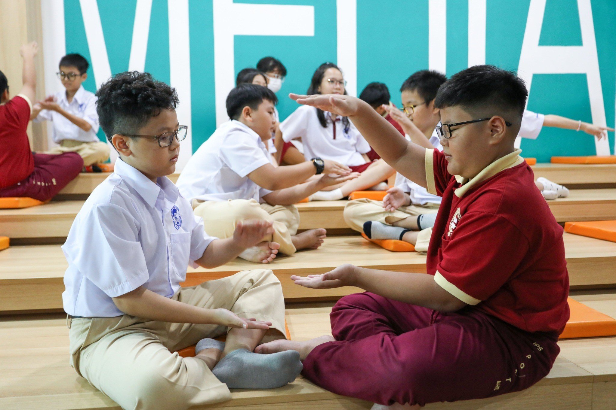 Hai nam sinh (hai học sinh nam) đang chơi trò vỗ tay; mặc áo sơ mi trắng/áo polo đỏ, quần kaki/đỏ, đeo kính; ngồi trên bậc thang gỗ có đệm cam trong không gian học tập sáng sủa.