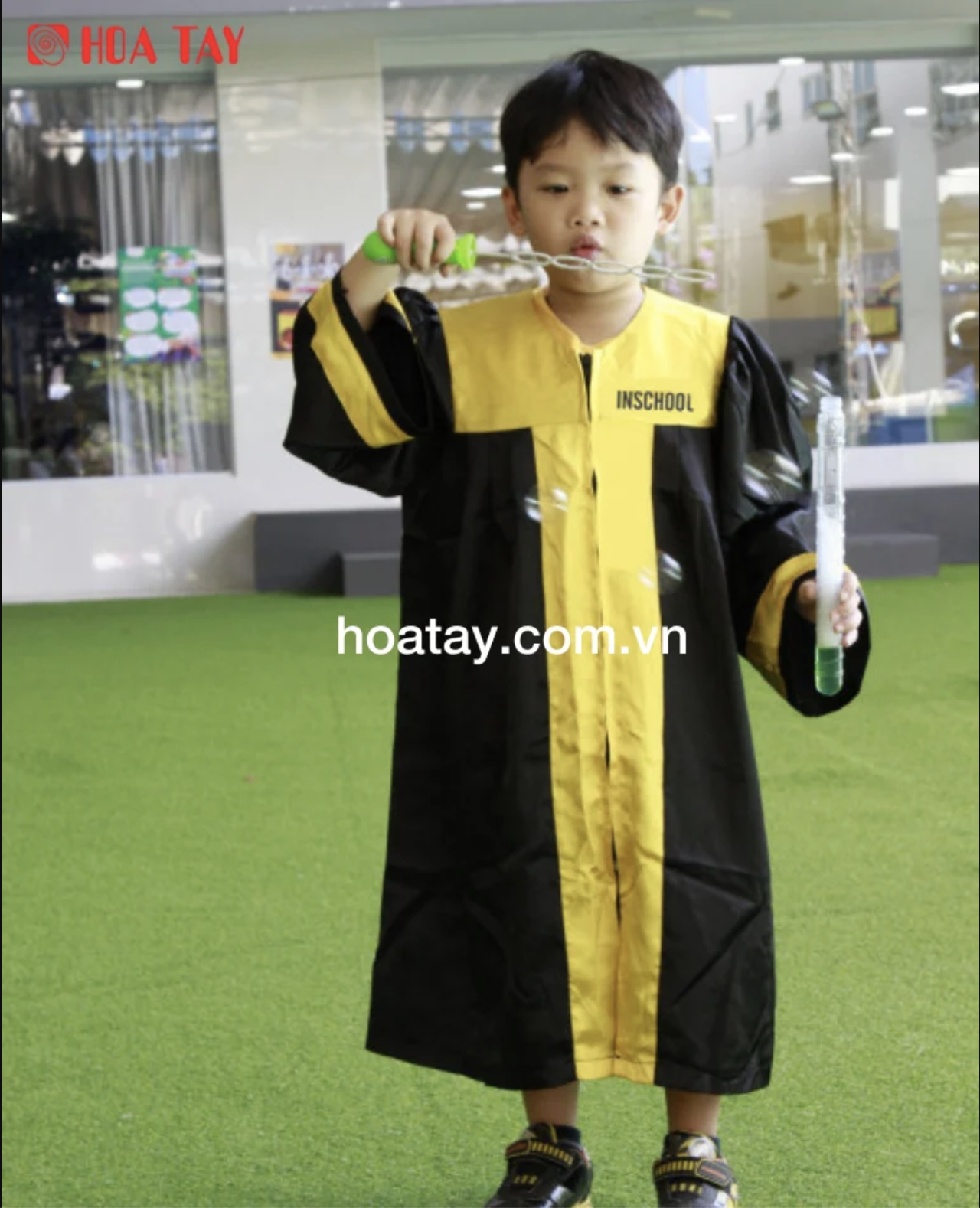 Một bé trai đang thổi bong bóng, mặc áo cử nhân (áo tốt nghiệp) màu đen phối vàng, tay áo rộng, cổ tròn, vải polyester bóng, chụp ngoài trời trên sân cỏ nhân tạo.