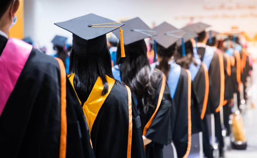 Hàng dài sinh viên tốt nghiệp đứng quay lưng, đội mũ cử nhân (tứ giác) màu đen có tua vàng, mặc áo toga đen với dải viền vàng và cam, trong hội trường sáng sủa.