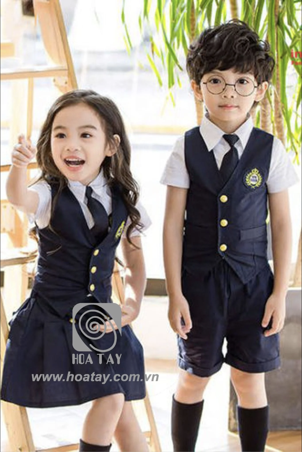 Hai trẻ em (một trai, một gái) đang đứng, mặc đồng phục học sinh kiểu vest (áo gile) màu xanh navy, sơ mi trắng tay ngắn, cà vạt đen, huy hiệu thêu ngực, cúc vàng, chân váy xếp ly và quần short, ánh s