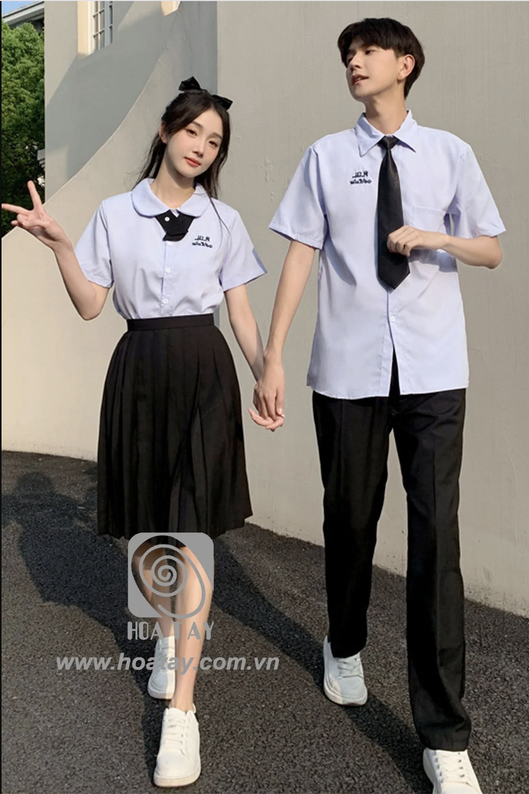 Một đôi nam nữ (hai người) đang đi bộ, mặc đồng phục học sinh kiểu Nhật (seifuku): áo sơ mi trắng tay ngắn, nữ mặc chân váy xếp ly (pleated skirt) đen, nam mặc quần tây đen, thắt cà vạt, ngoài trời nắ