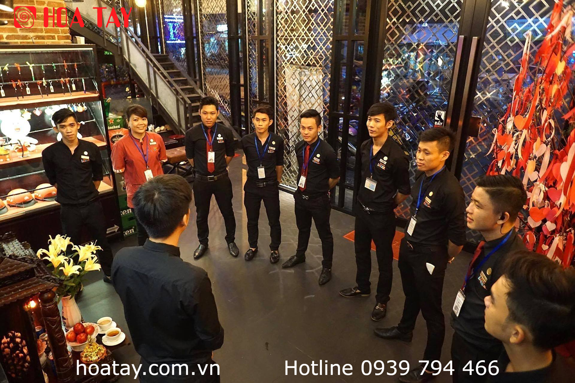 Nhóm 9 nhân viên (team) đang họp đầu ca; mặc sơ mi đen (black shirt), quần tây (trousers), đeo thẻ tên (ID badge) và cà vạt đỏ; không gian nhà hàng hiện đại, ánh sáng vàng ấm.