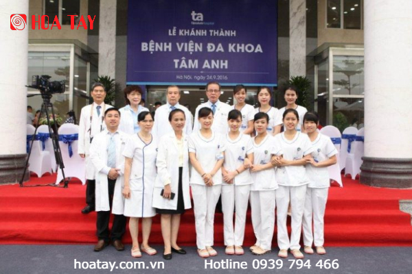 Nhóm khoảng 15 nhân viên y tế đứng trên thảm đỏ trước biển hiệu 'Lễ khánh thành Bệnh viện đa khoa Tâm Anh'. Họ mặc áo blouse trắng, áo choàng dài, và đồng phục y tá trắng. Phía sau là ghế bọc trắng và