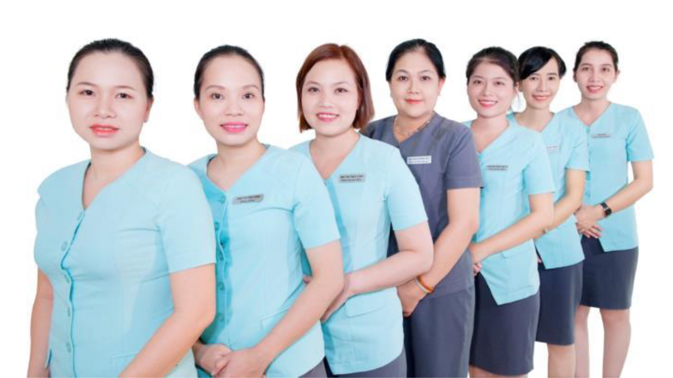 7 nữ nhân viên y tế đứng thành hàng -> mặc đồng phục -> áo polo cổ V màu xanh da trời nhạt, một người mặc áo xám, quần tây màu xám đậm, tên hiệu trên ngực trái -> phông nền trắng, ánh sáng studio đều