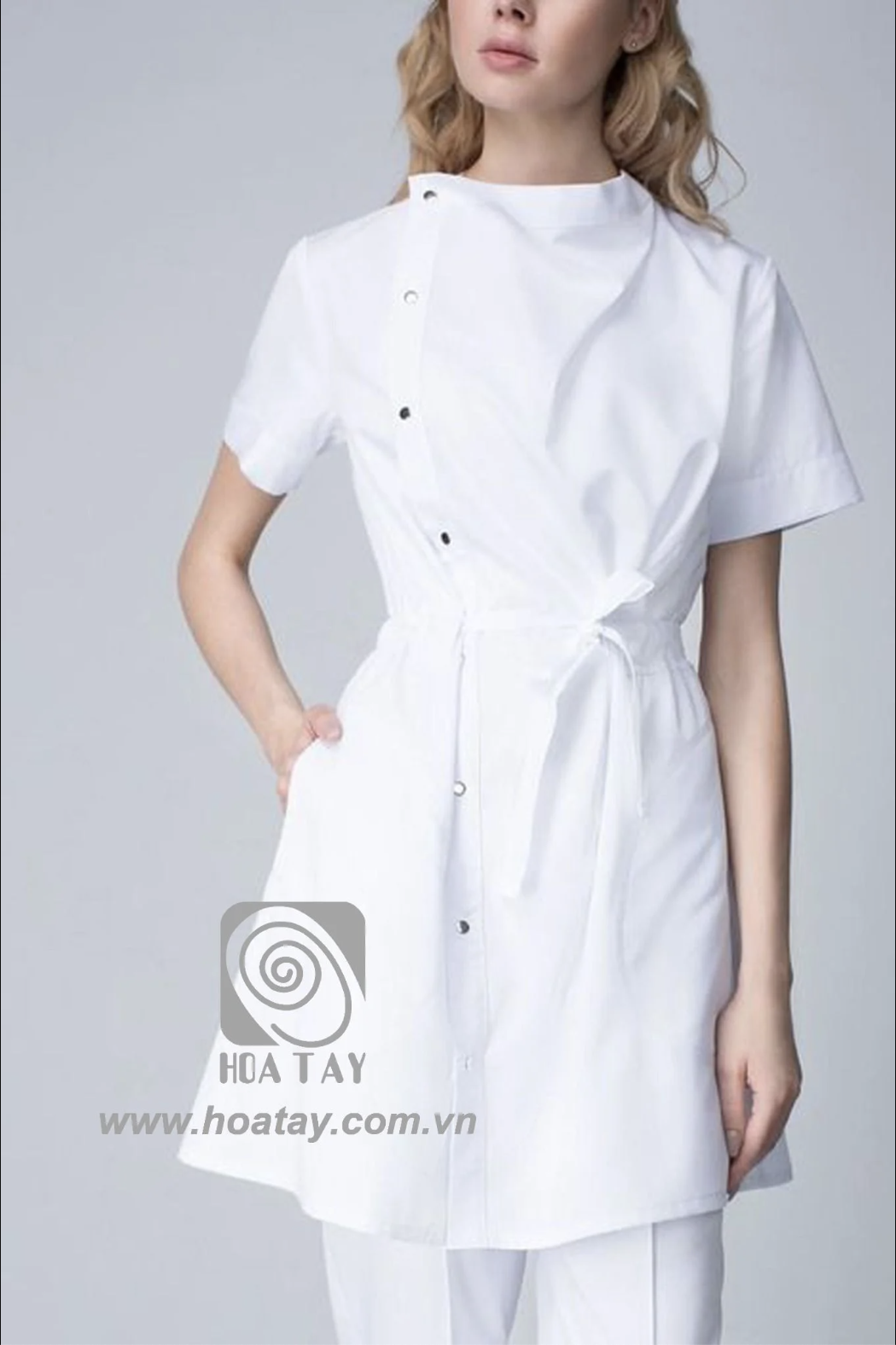 Một nữ nhân viên y tế mặc áo blouse trắng cổ chéo (asymmetrical neckline) với hàng khuy bấm (snap buttons) dọc thân, chất liệu vải cotton phẳng, tay ngắn, thắt đai eo, nền xám trung tính, ánh sáng stu