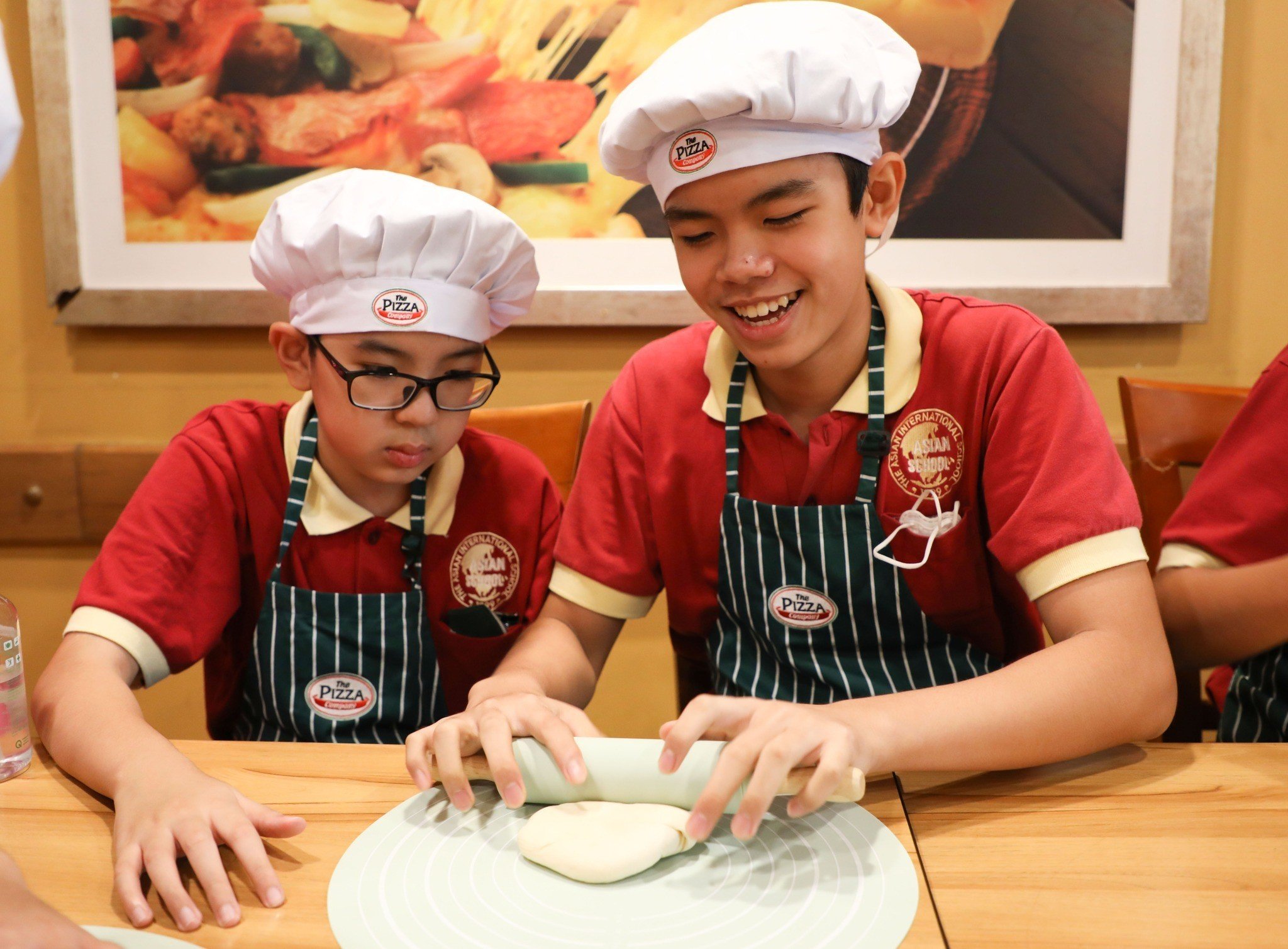 Hai cậu bé (two boys) đang cán bột (rolling dough) đội mũ đầu bếp (chef hats) màu trắng, mặc áo polo đỏ phối tạp dề kẻ sọc (striped aprons) trong nhà hàng, ánh sáng vàng ấm áp.