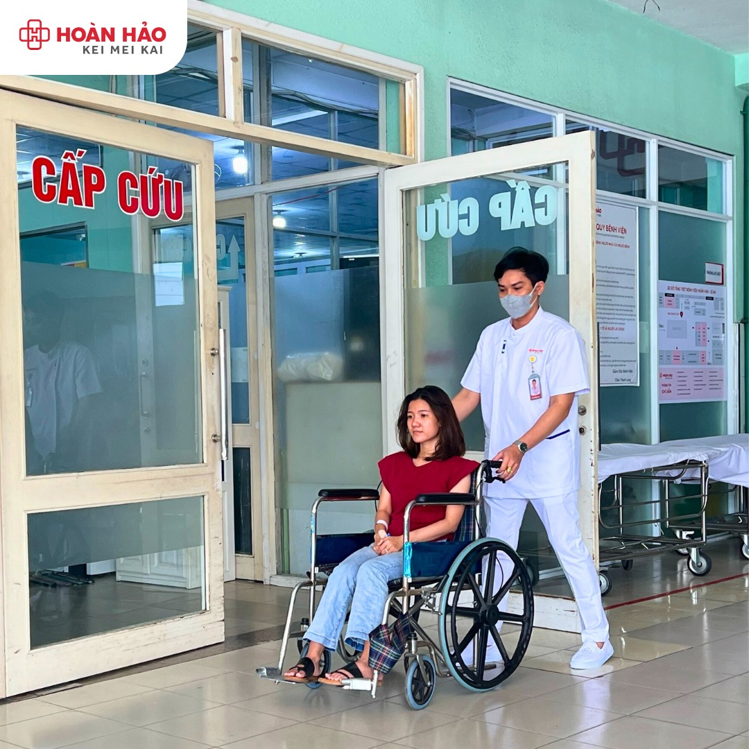 Một nam điều dưỡng đẩy một nữ bệnh nhân trên xe lăn (ghế có bánh xe) qua cửa phòng cấp cứu; điều dưỡng mặc áo blouse trắng cổ chữ V, quần dài trắng, đeo khẩu trang y tế; bối cảnh bệnh viện, ánh sáng t