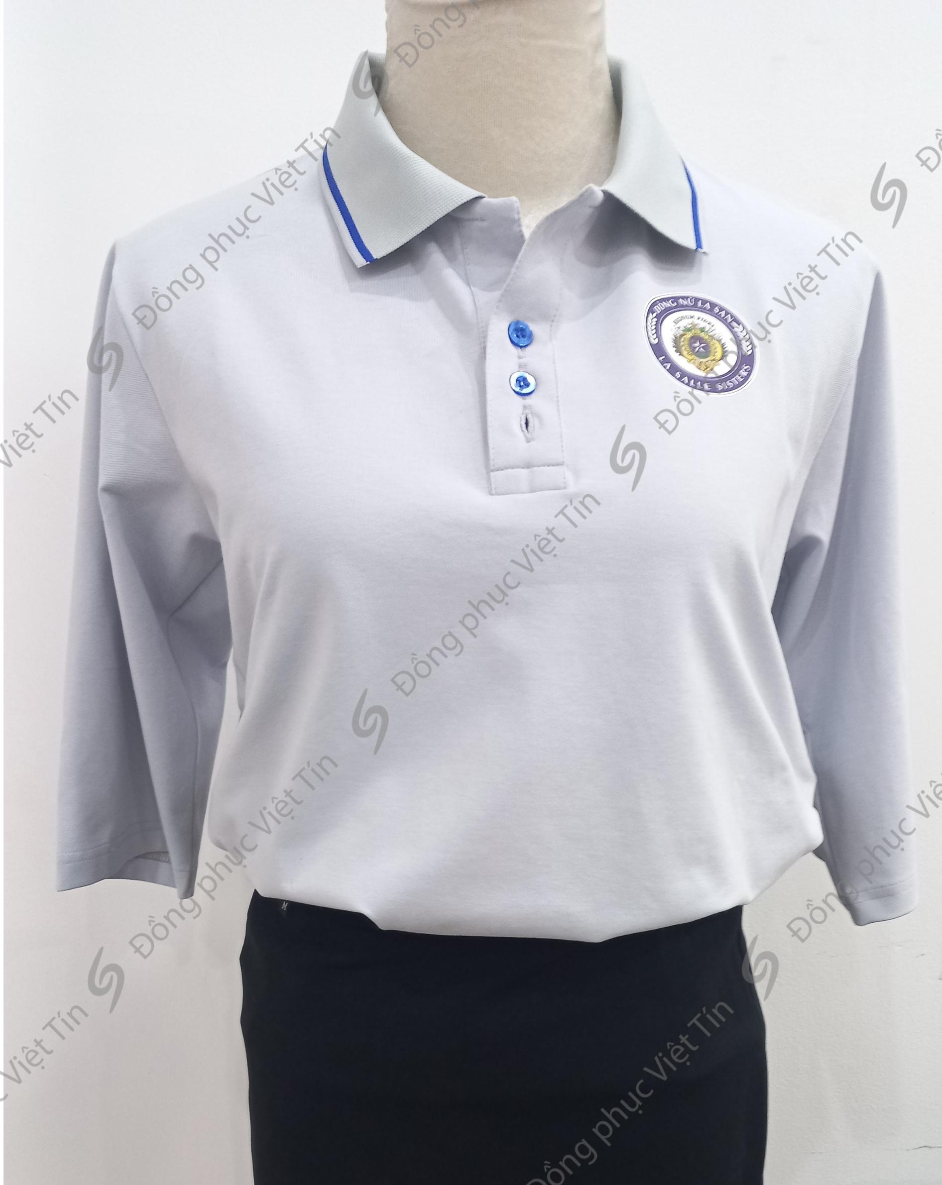 Áo polo màu xám nhạt tay lỡ, cổ áo có viền xanh, chất liệu Cá sấu Mỹ, cúc áo xanh, logo trường La Salle Sisters trên ngực trái, mặc trên ma-nơ-canh, nền trắng.
