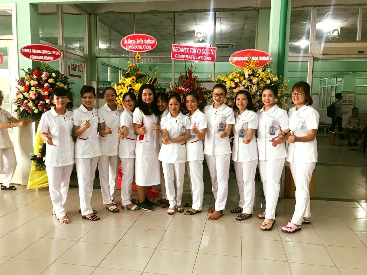 Nhóm 12 nữ nhân viên y tế mặc đồng phục trắng (áo cổ bẻ, quần dài) đứng thành hàng, làm dấu tay, trước các lẵng hoa chúc mừng và biển tên công ty trong sảnh bệnh viện có sàn gạch sáng.