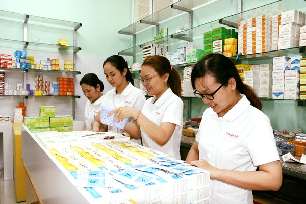 Bốn nữ nhân viên trong áo polo trắng đứng sau quầy kính, kiểm tra các hộp thuốc màu xanh và vàng; kệ kính phía sau chứa đầy hộp thuốc, ánh sáng trắng sáng sủa.