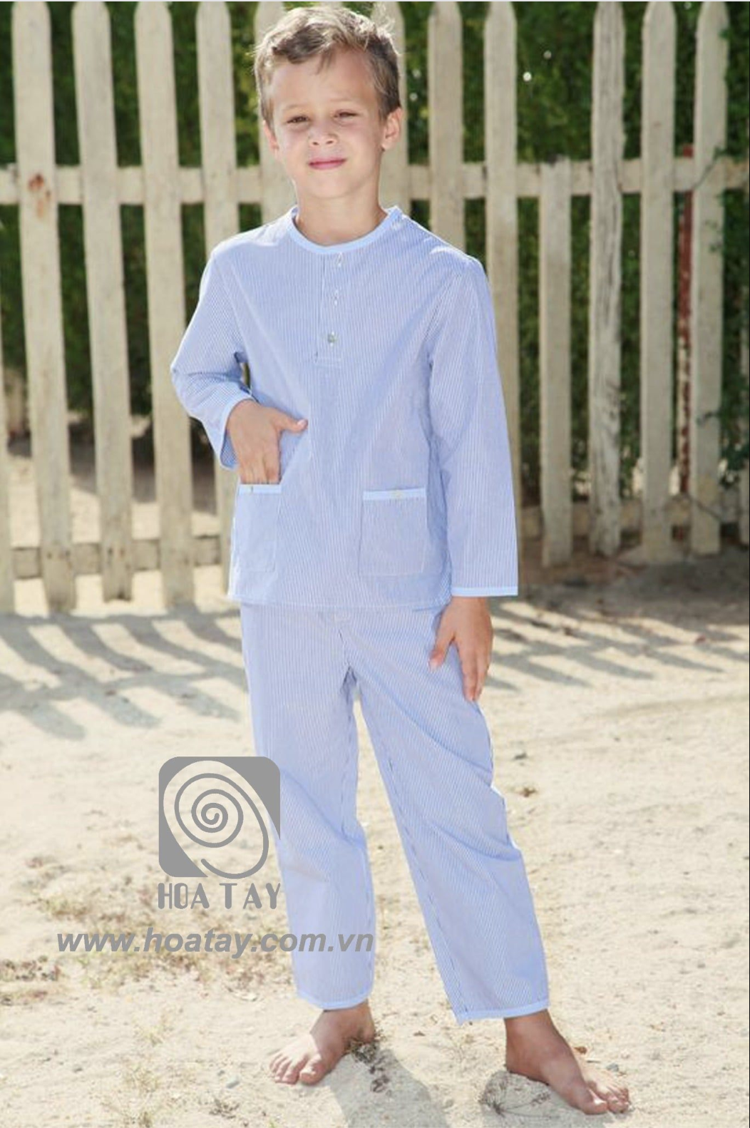 Một bé trai đang đứng, mặc bộ đồ ngủ (pajamas) kẻ sọc xanh trắng, cổ tròn (crew neck), tay dài, có hai túi trước, chất liệu cotton mỏng, bối cảnh ngoài trời cạnh hàng rào gỗ, ánh sáng tự nhiên.
