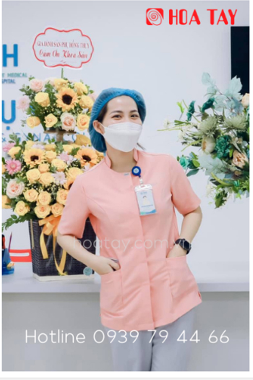 Một nữ nhân viên y tế (điều dưỡng) đang đứng, mặc áo blouse cổ tròn (cổ tàu) màu hồng phấn, tay ngắn, có hai túi trước, đội mũ bao tóc màu xanh dương, đeo khẩu trang trắng, bối cảnh bệnh viện với lẵng