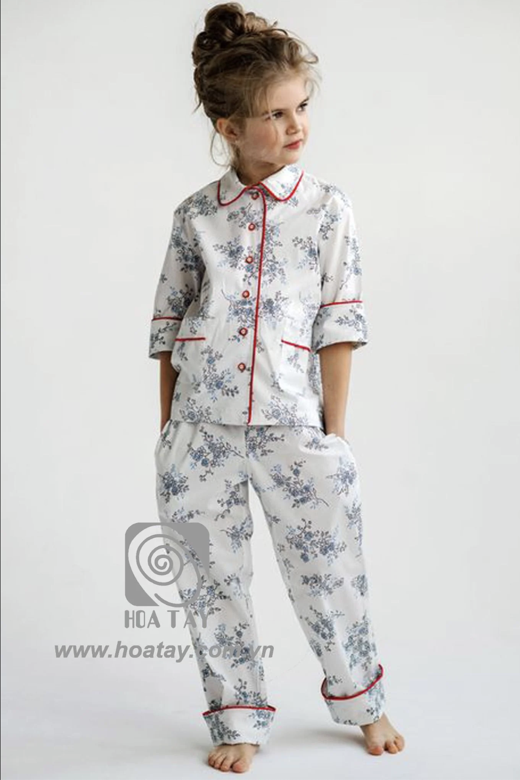 Một bé gái mặc bộ đồ ngủ vải cotton trắng in hoa xanh, cổ bẻ viền đỏ, tạo cảm giác thoải mái và an toàn