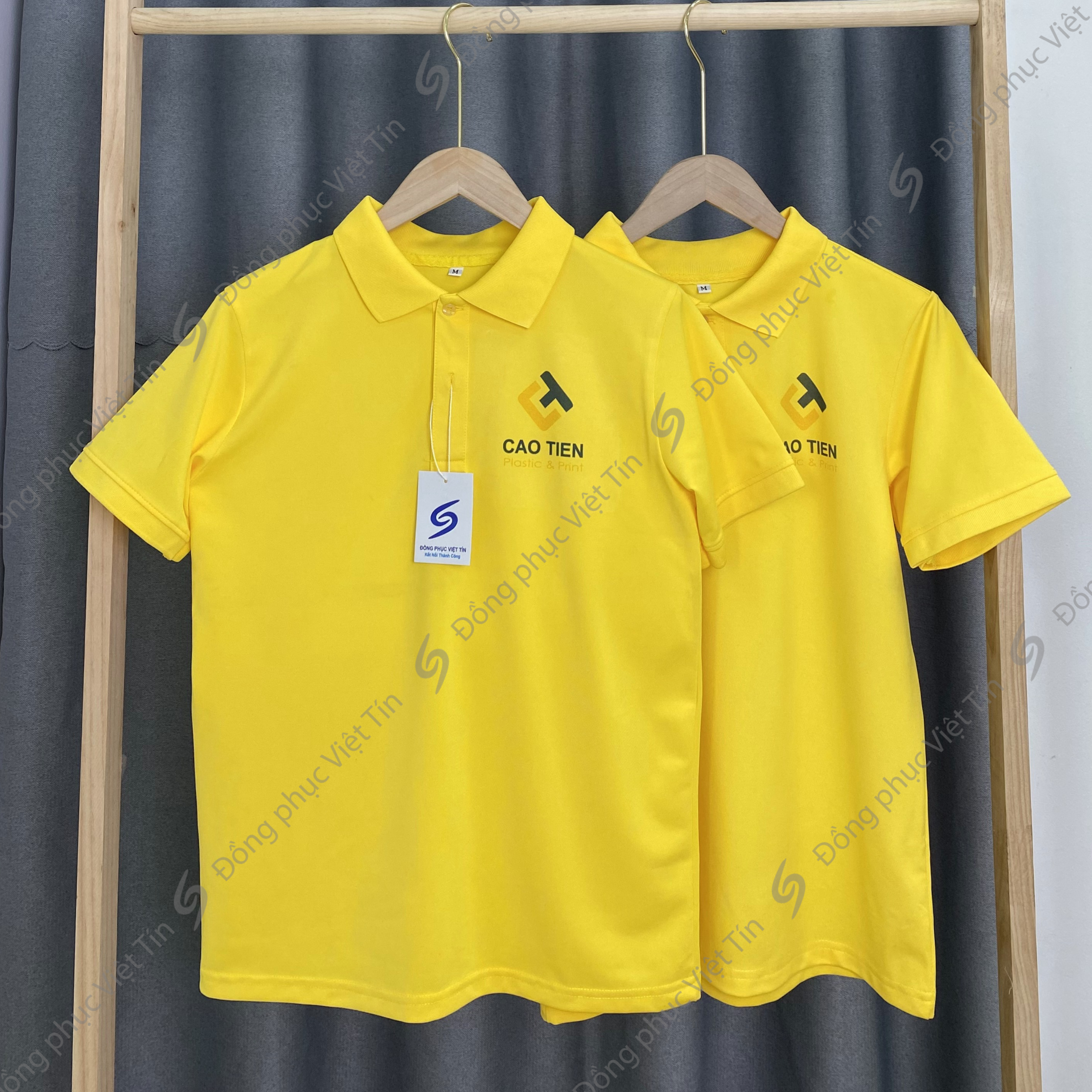 Hai chiếc áo polo màu vàng tươi có cổ, chất liệu Poly tiêu chuẩn, in logo 'CAO TIEN' màu đen, được treo trên móc gỗ trên giá treo quần áo với phông nền rèm xám.