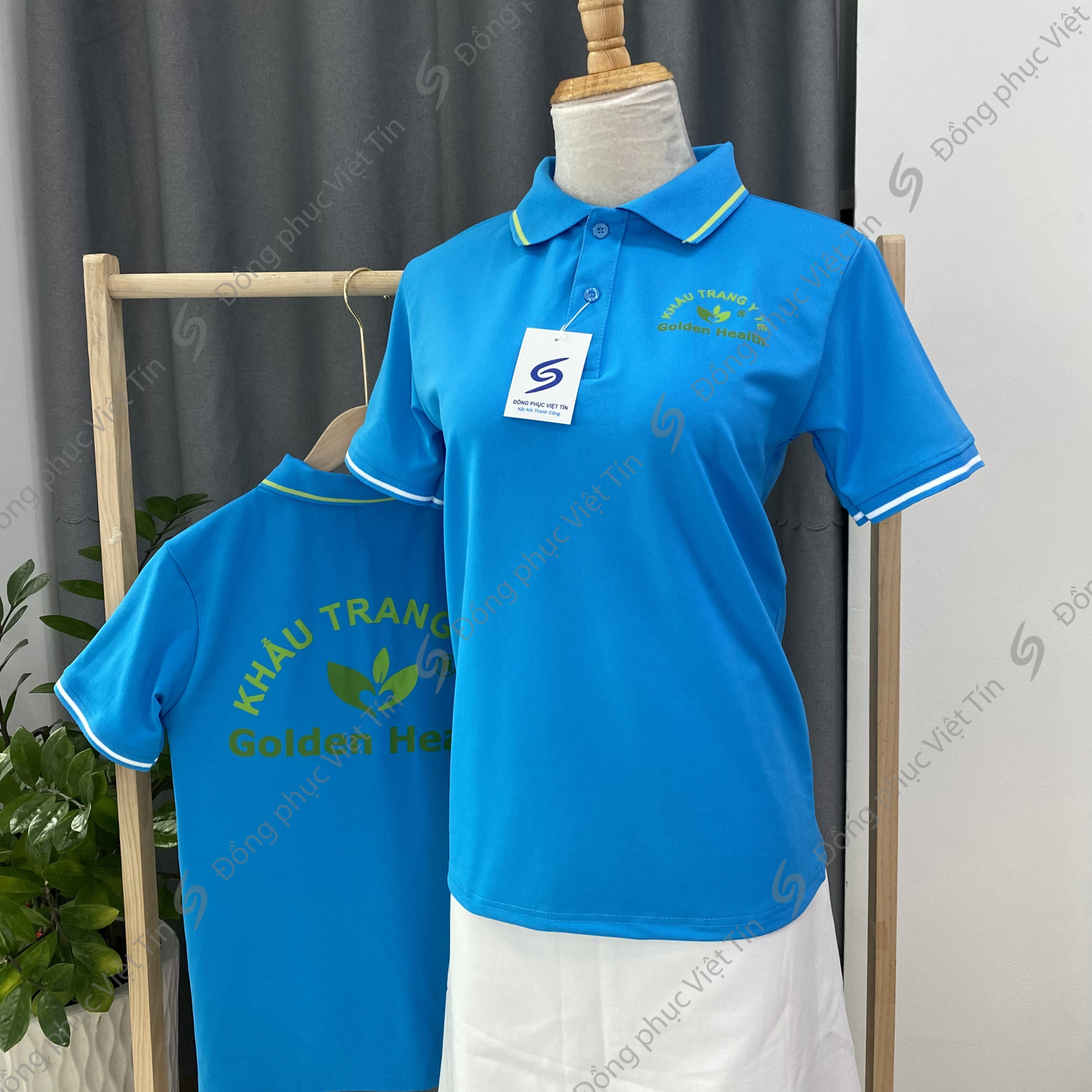 Áo polo màu xanh dương tươi sáng với logo 'Golden Health' màu xanh lá trước sau, chất liệu Poly tiêu chuẩn, được trưng bày trên ma-nơ-canh và móc treo gỗ trong không gian studio tối giản.
