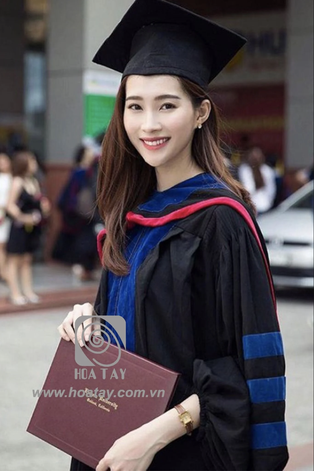 Một nữ sinh viên tốt nghiệp đang mỉm cười, mặc áo cử nhân (áo lễ phục) màu đen phối dải màu xanh dương, đội mũ mortarboard (mũ tốt nghiệp vuông) màu đen, cầm bằng tốt nghiệp, bối cảnh khuôn viên trườn