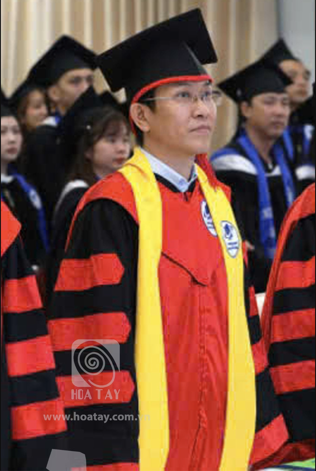Một nam sinh viên đội mũ cử nhân (mũ vuông) đang đứng dự lễ tốt nghiệp; mặc áo thụng đỏ phối đen với dải khăn choàng vàng, chất liệu vải satin bóng, chi tiết tay áo xếp ly, bối cảnh hội trường đông ng