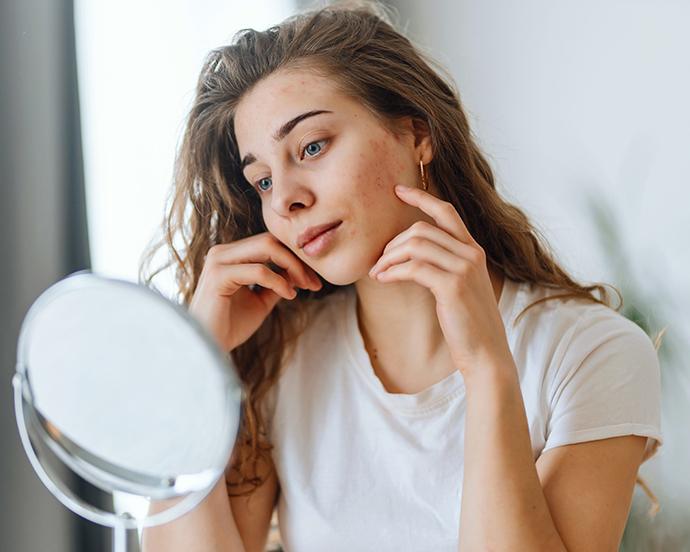 Cô gái trẻ soi gương, kiểm tra làn da đang có vấn đề, minh họa cho tầm quan trọng của quy trình chăm sóc da cơ bản.