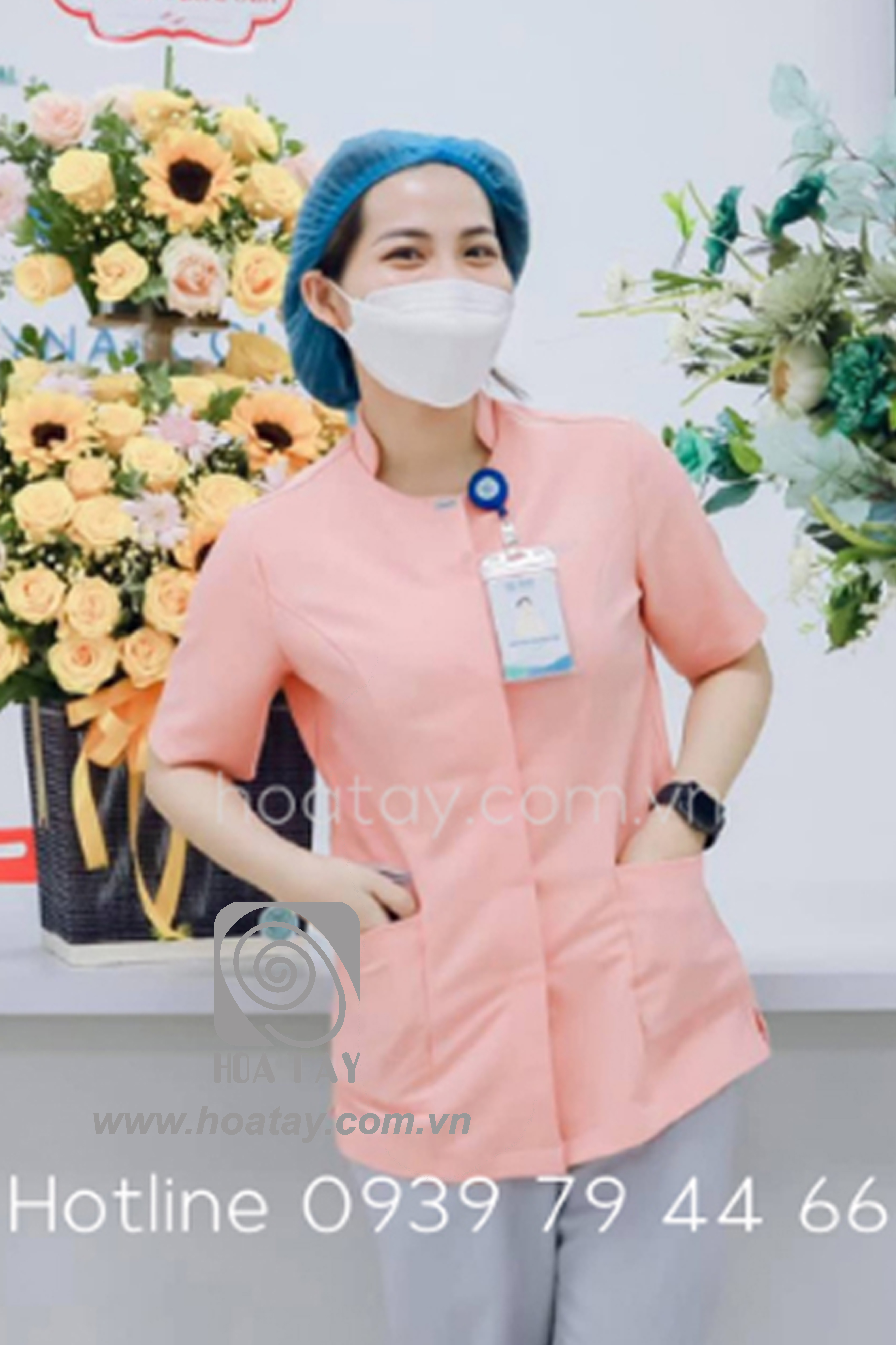 Một nữ nhân viên y tế đang đứng, mặc áo blouse cổ tròn (cổ tàu) màu hồng phấn, tay ngắn, chất liệu vải phẳng mịn, có hai túi hông lớn, đeo thẻ tên và mũ trùm đầu y tế màu xanh, bối cảnh trong nhà với