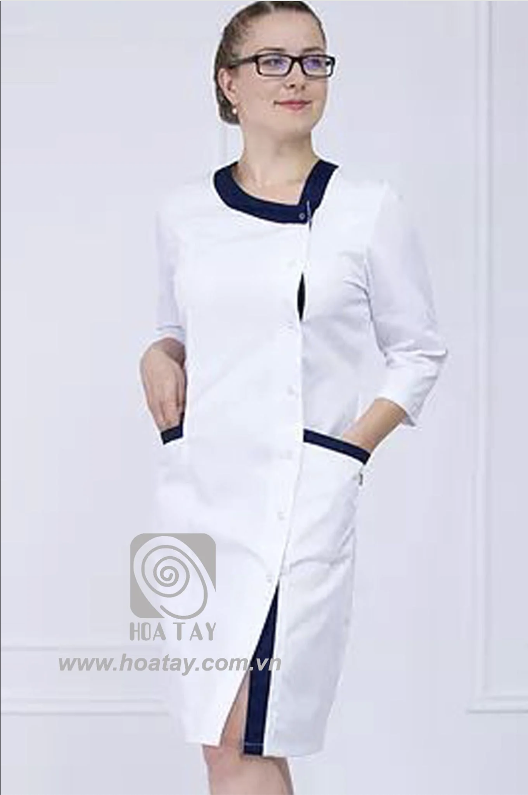 Một nữ nhân viên y tế đeo kính đang đứng; áo blouse trắng cổ tàu phối viền xanh navy, tay lỡ, cúc bấm ẩn, hai túi hông có viền xanh, chất vải cotton phẳng phiu; phông nền trắng, ánh sáng studio đều.