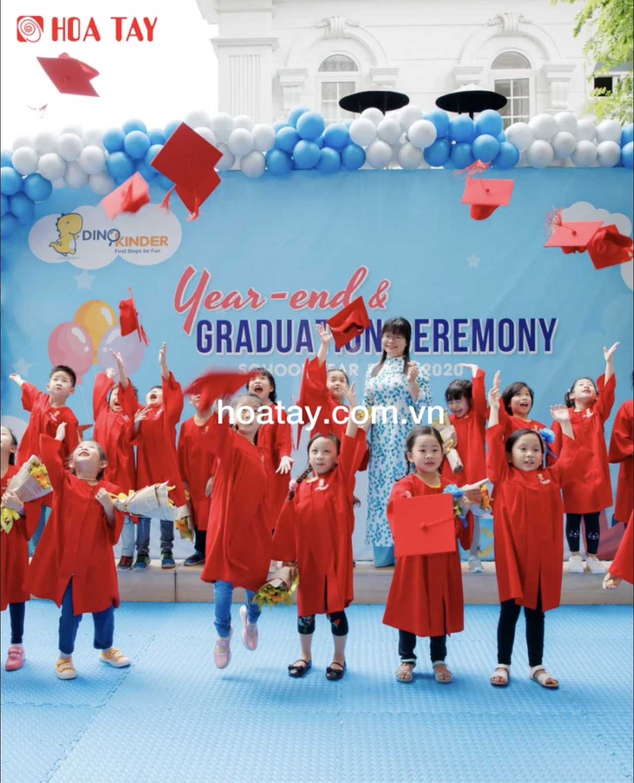 Nhóm học sinh mầm non (khoảng 15 trẻ) đang tung mũ cử nhân (mũ tốt nghiệp) lên không trung; mặc áo lễ phục (áo choàng) đỏ, chất liệu vải trơn, phom rộng, tay dài; bối cảnh sân khấu ngoài trời trang tr
