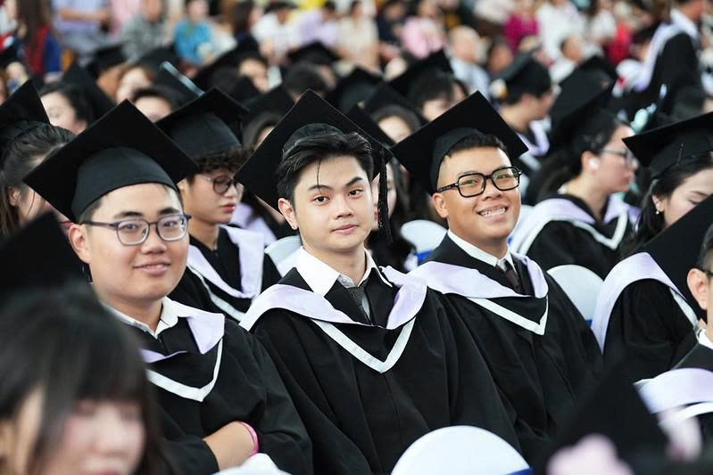 Ba nam sinh (ba người) đang ngồi dự lễ tốt nghiệp, mặc áo cử nhân (áo choàng đen) với dải khăn quàng cổ (vải satin màu trắng/tím nhạt) và đội mũ tốt nghiệp (mũ vuông có tua rua), bối cảnh hội trường đ