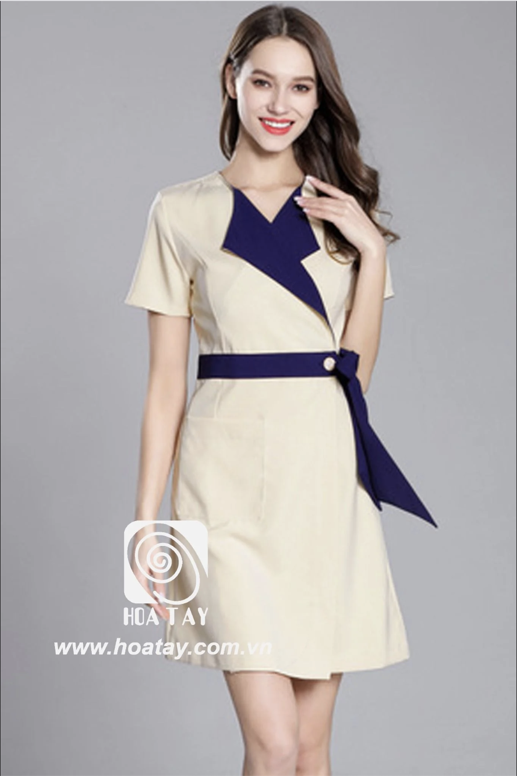 Một nữ nhân viên đang đứng, mặc váy đồng phục (đầm liền) màu kem phối xanh navy, cổ chữ V (cổ xẻ), thắt đai eo (dây buộc) có cúc, tay ngắn, chất vải phẳng mịn, phông nền xám, ánh sáng studio.