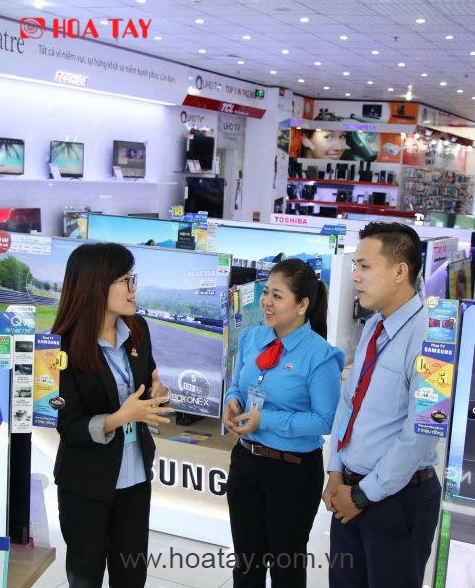 Ba nhân viên (một nữ mặc vest đen, hai nhân viên mặc sơ mi xanh) đang trao đổi công việc; trang phục công sở chuyên nghiệp, bảng tên đeo cổ, không gian cửa hàng điện máy sáng trưng với nhiều màn hình
