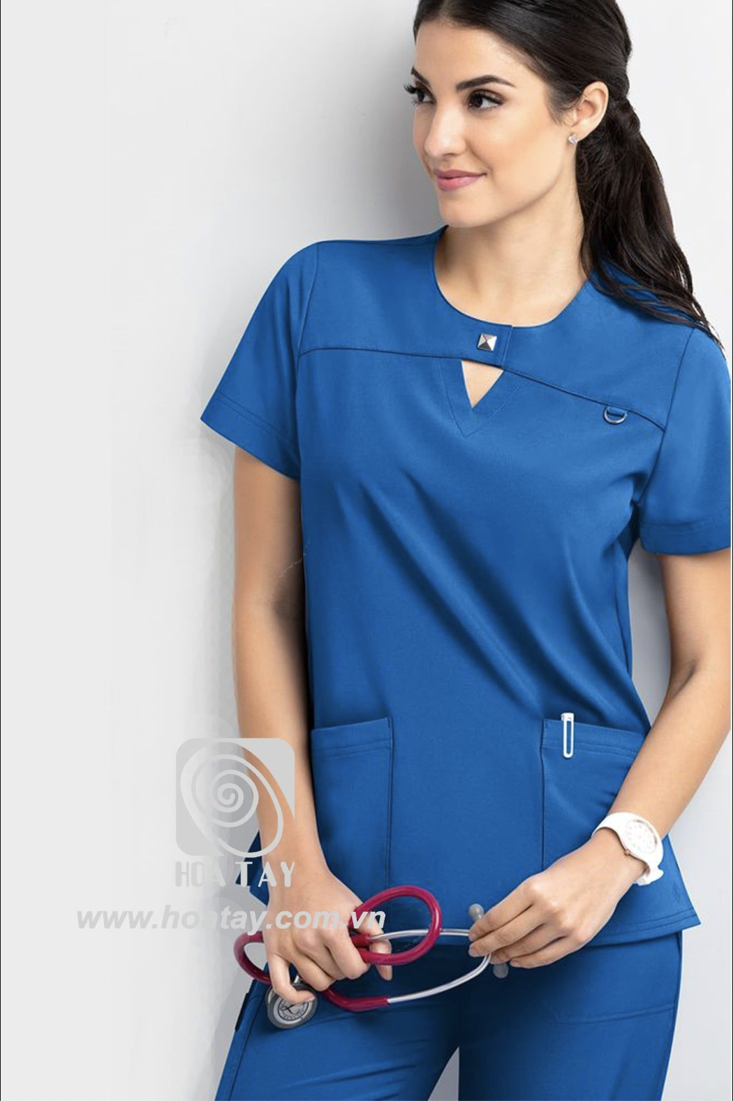 Một nữ nhân viên y tế đang đứng, mặc áo blouse cổ chữ V (v-neck) màu xanh dương, chất liệu vải co giãn, có chi tiết đinh tán kim loại ở cổ và túi trước, phông nền studio sáng.