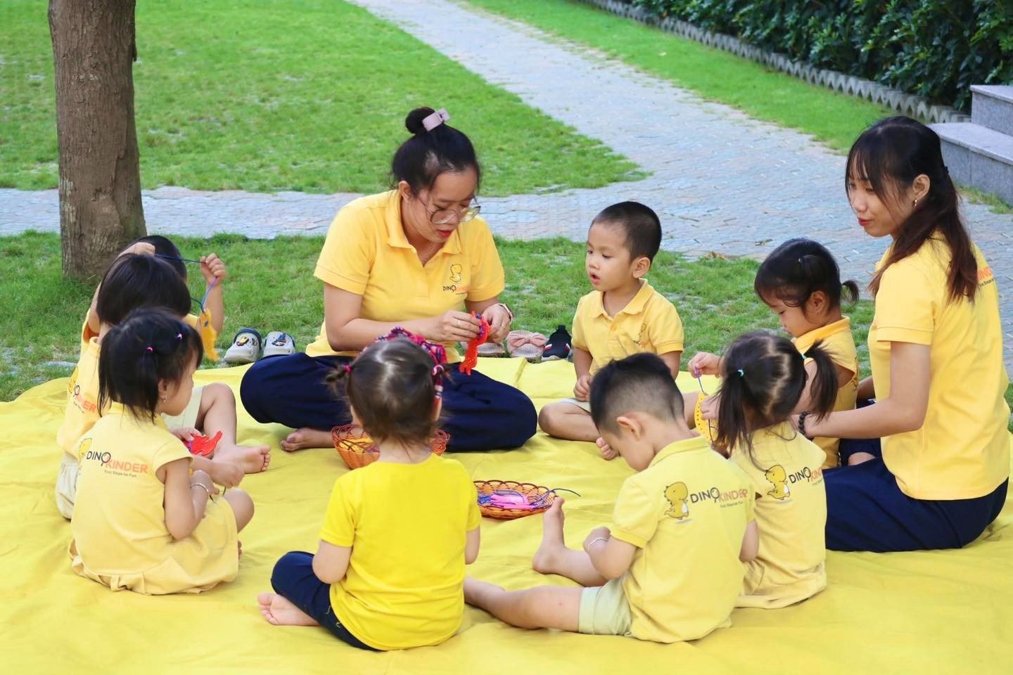 Một nhóm giáo viên và trẻ mầm non (9 người) đang ngồi vòng tròn trên thảm vàng ngoài trời; mặc áo polo đồng phục màu vàng, quần tối màu; không gian công viên, ánh sáng tự nhiên ban ngày.
