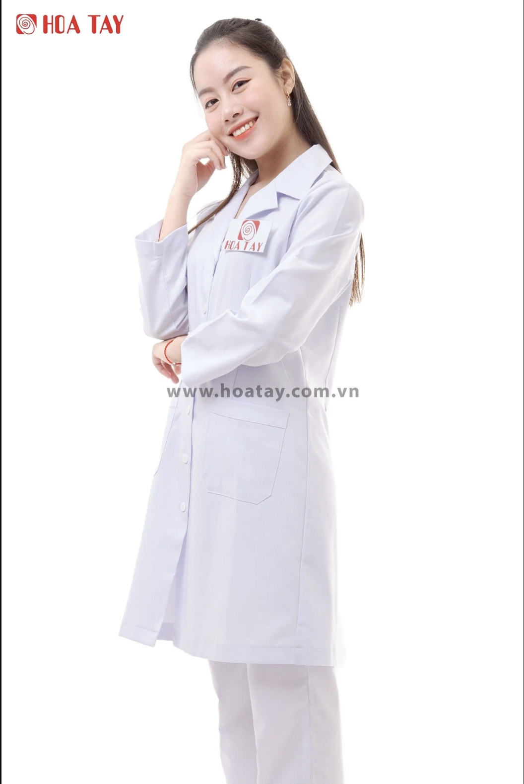 Một nữ nhân viên y tế đang đứng mỉm cười, mặc áo blouse trắng (áo khoác phòng thí nghiệm) dài tay, cổ bẻ (cổ ve), chất liệu vải cotton phẳng phiu, có túi vuông phía trước, nền trắng sáng studio.