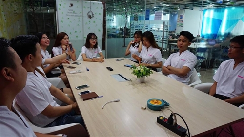 Nhóm sinh viên (hơn 30 người) đang tung mũ cử nhân (mũ tốt nghiệp vuông) lên không trung; mặc áo lễ phục (áo choàng đen) dáng rộng, vải polyester, cổ chữ V; chụp ngoài trời tại sân thể thao, ánh sáng