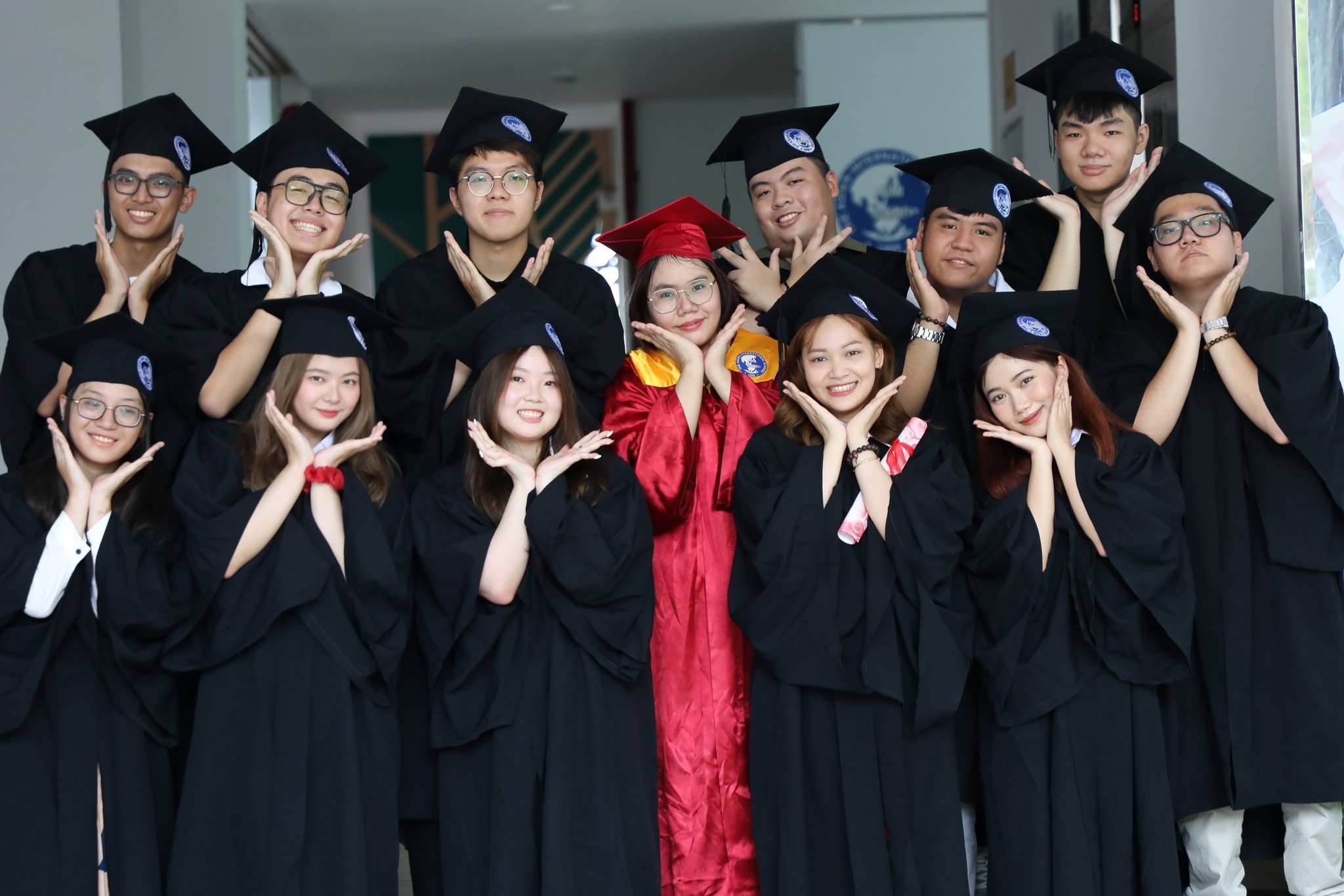 11 sinh viên tốt nghiệp (tân cử nhân) đang tạo dáng tay hình trái tim, mặc áo choàng đen (áo cử nhân) và mũ vuông (nón tốt nghiệp), một người mặc áo choàng đỏ, bối cảnh hành lang trường học, ánh sáng