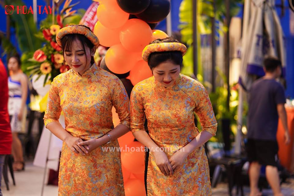 Hai phụ nữ (hai người) đang cúi chào; mặc áo dài truyền thống (áo dài cổ cao) màu vàng cam, họa tiết hoa văn chìm, tay lỡ, đội mấn (khăn vấn) cùng bộ; bối cảnh sự kiện ngoài trời với bóng bay trang tr