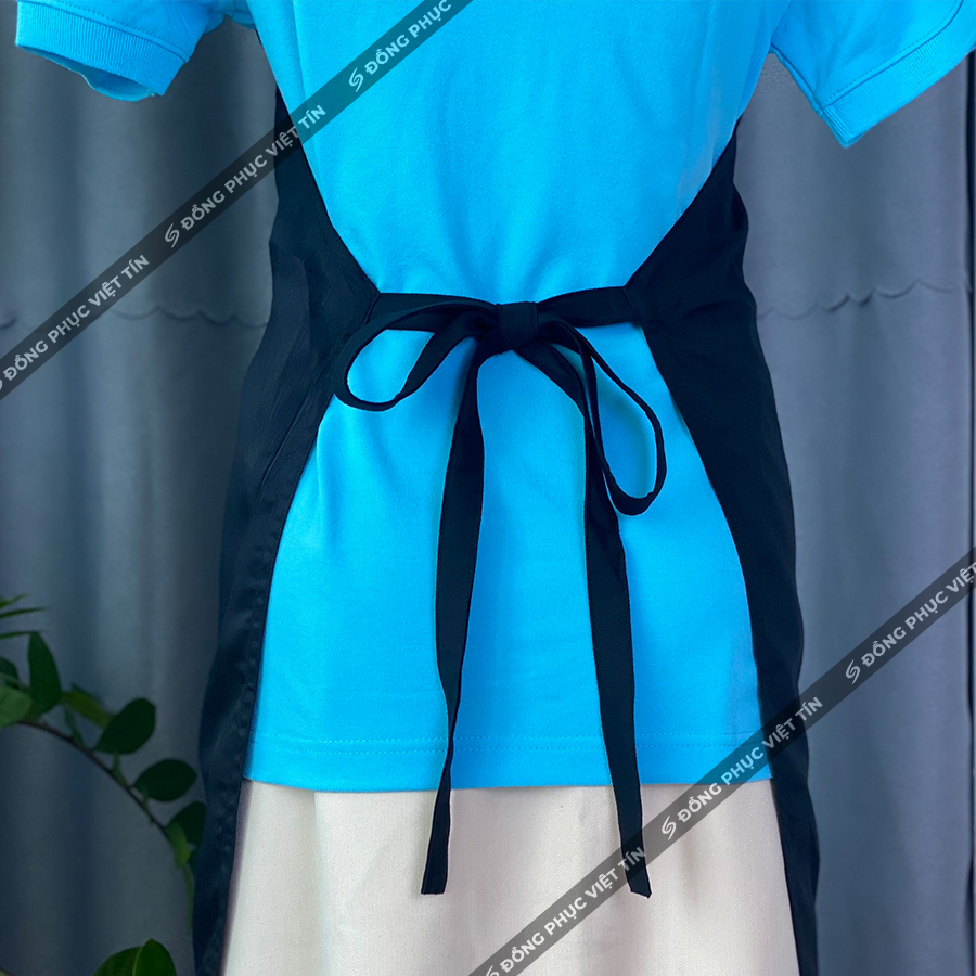 Hình ảnh chụp phía sau một người mặc áo thun màu xanh dương kết hợp với tạp dề màu đen thắt nơ ở eo, trên nền vải xám và có chèn logo thương hiệu chéo.