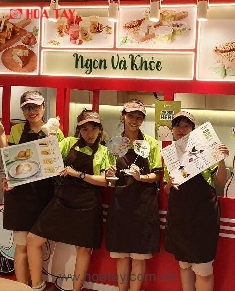 Bốn nhân viên nữ đang đứng, mặc áo polo xanh lá phối tạp dề đen (yếm), đội mũ lưỡi trai cùng màu, cầm thực đơn và bảng hiệu nhỏ, bối cảnh quầy hàng đồ uống có đèn LED rực rỡ.
