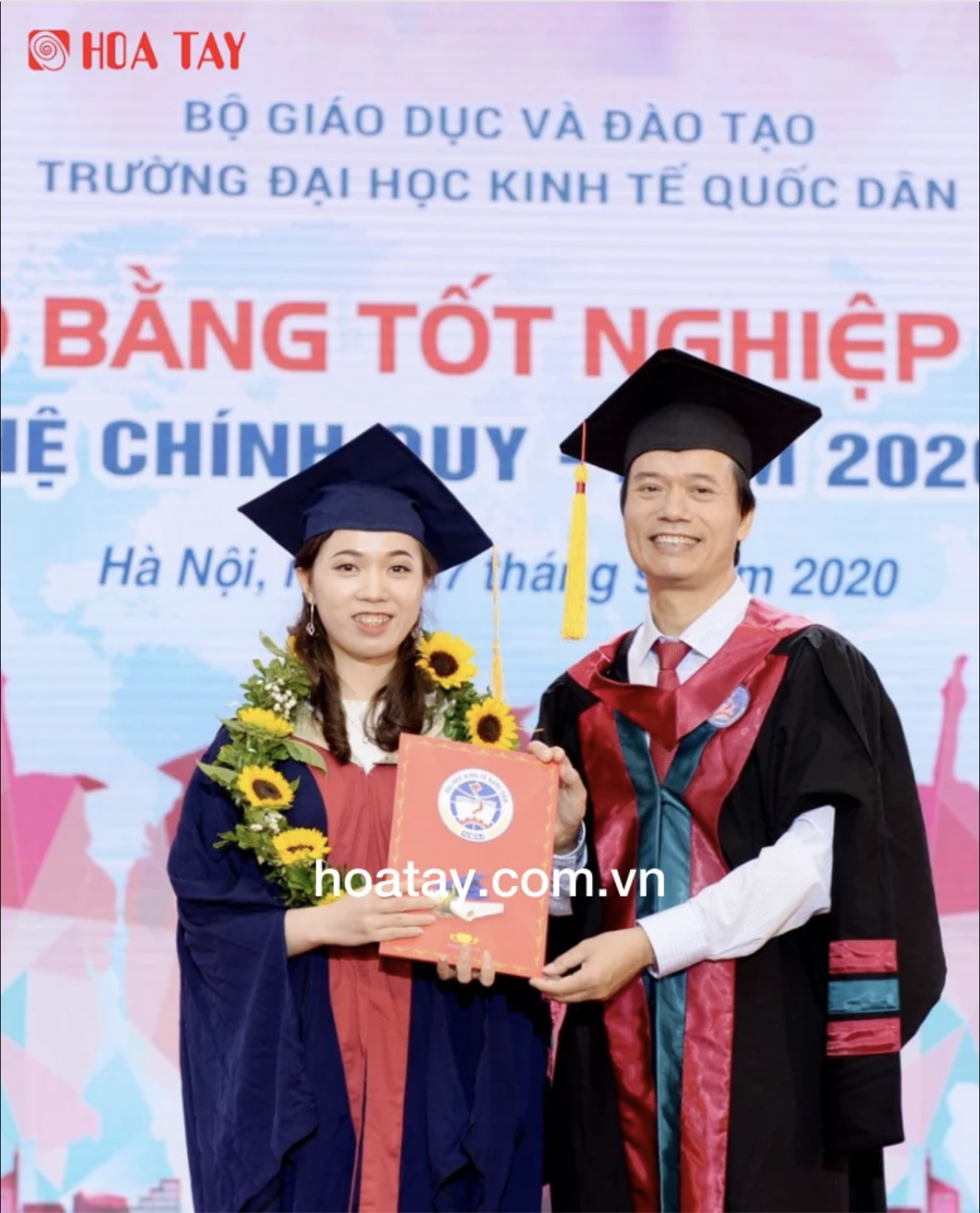 Hai người (một sinh viên, một giảng viên) đang trao bằng tốt nghiệp; mặc áo cử nhân (áo thụng) màu xanh đen, khăn choàng đỏ, mũ vuông (nón tốt nghiệp) có tua rua, vòng hoa hướng dương; bối cảnh sân kh