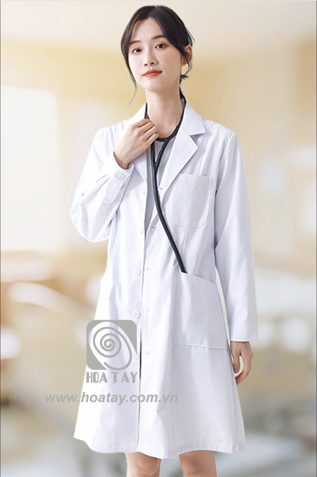 Một nữ bác sĩ đang đứng, mặc áo blouse trắng (áo khoác phòng thí nghiệm) dài tay, cổ bẻ (cổ ve), chất liệu vải cotton phẳng, có túi ngực và túi hông, đeo ống nghe, bối cảnh bệnh viện mờ, ánh sáng dịu.