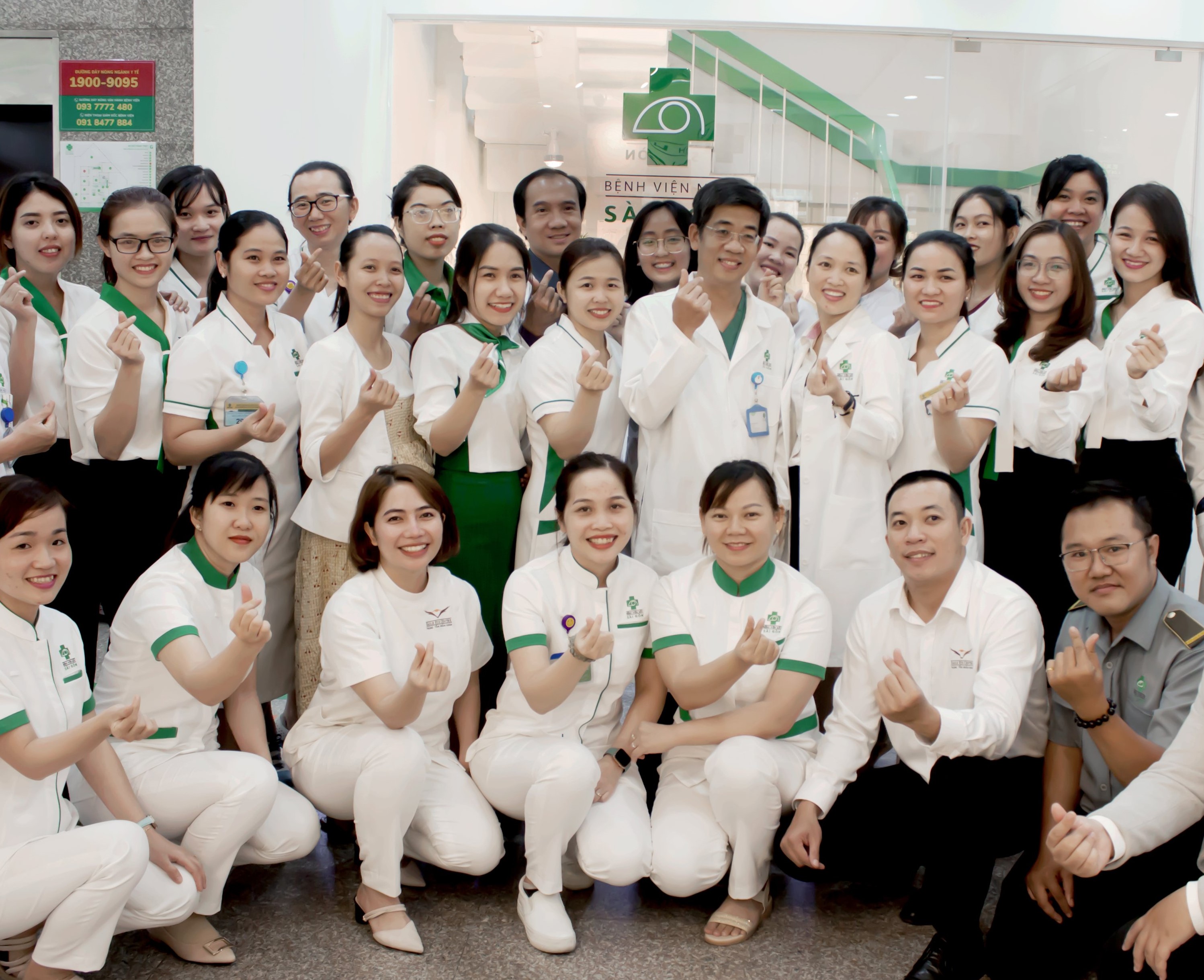 Nhóm khoảng 25 nhân viên y tế đứng và quỳ trong sảnh bệnh viện, mặc đồng phục trắng với viền cổ và tay áo màu xanh lá cây, một số người đeo bảng tên, tất cả cùng làm động tác tay 'yêu thương' hướng về