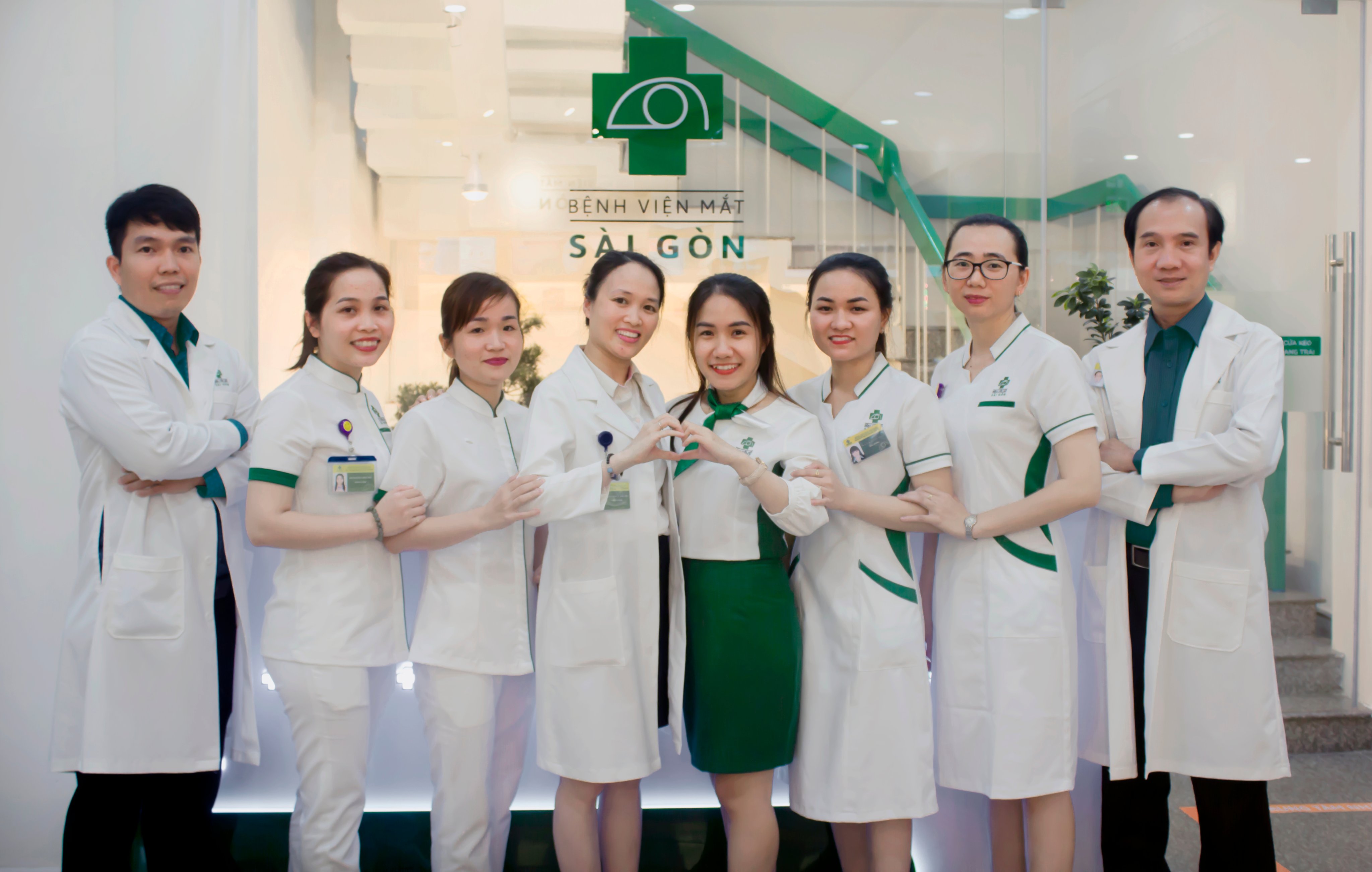 Bảy nhân viên y tế đứng thành hàng, tạo hình trái tim bằng tay. Áo blouse trắng có viền xanh lá, áo choàng trắng, đồng phục y tá. Nền là sảnh bệnh viện với logo 'Bệnh viện Mắt Sài Gòn' và cầu thang xa