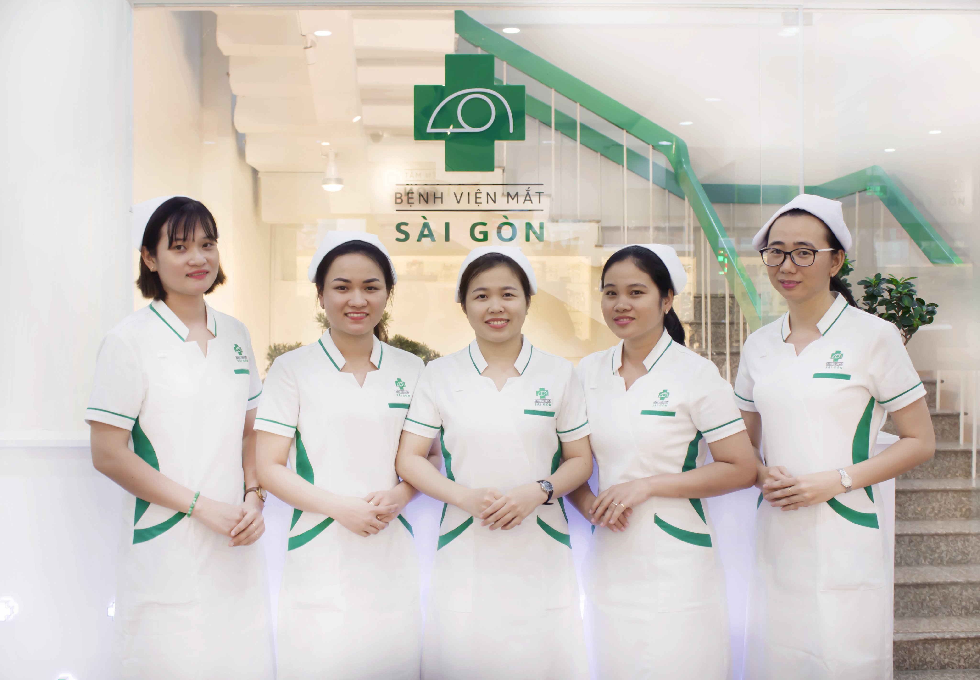 Năm nữ nhân viên y tế mặc đồng phục trắng có viền xanh lá, đeo mũ y tá, đứng trước logo Bệnh viện Mắt Sài Gòn