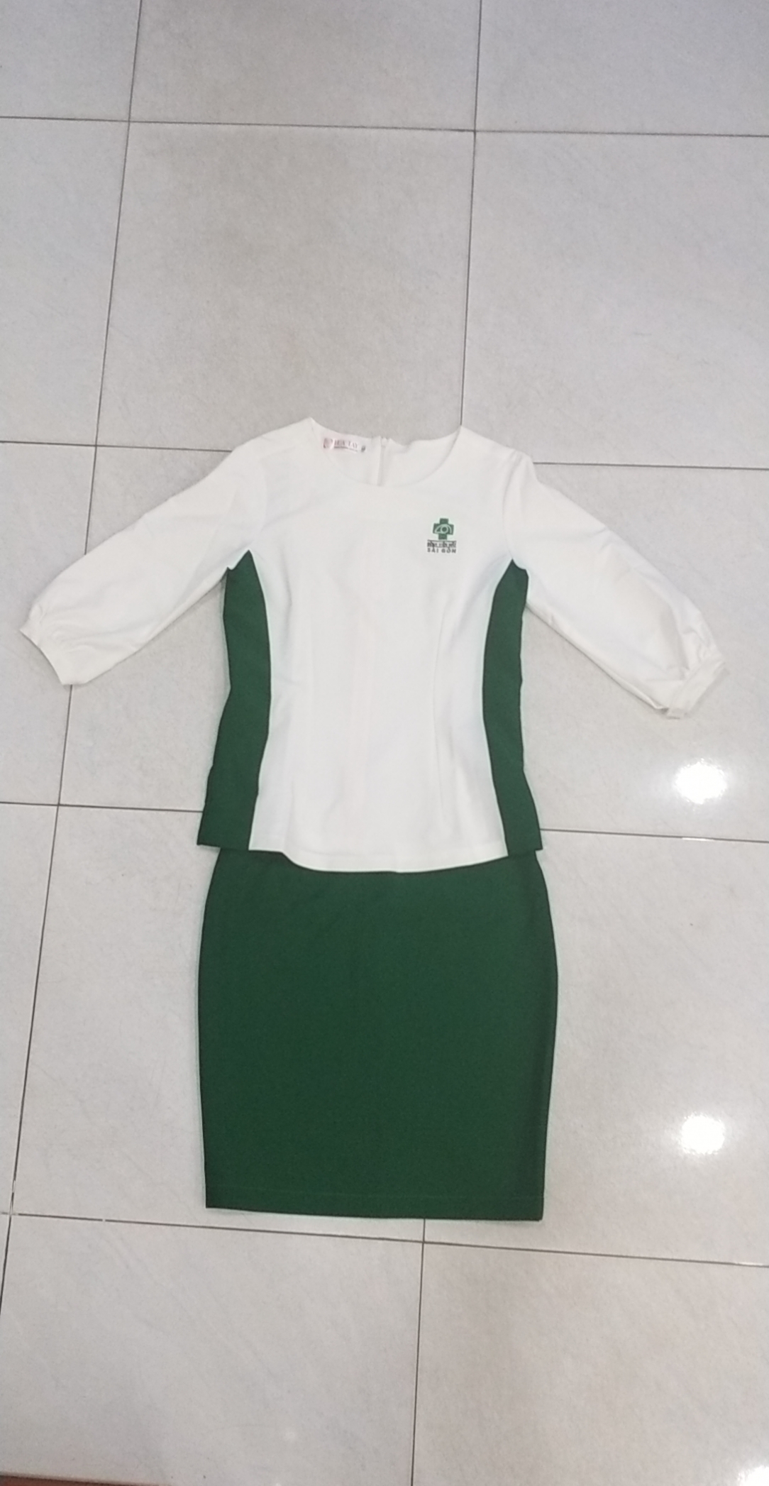Bộ đồng phục nữ gồm áo sơ mi trắng tay dài và chân váy bút chì xanh lá cây đậm, được trải phẳng trên sàn gạch men trắng. Áo có logo thêu màu xanh ở ngực trái, cổ tròn, và hai mảng vải xanh lá chạy dọc