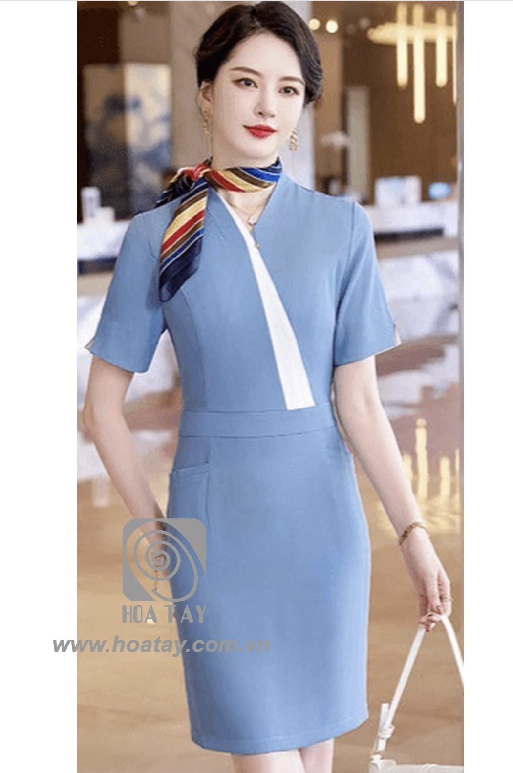 Một nữ nhân viên đang đứng, mặc váy đồng phục liền thân (dress) màu xanh dương nhạt, cổ chữ V phối vạt trắng, tay ngắn, thắt khăn lụa đa sắc, chất liệu vải phẳng mịn, bối cảnh văn phòng sang trọng, án