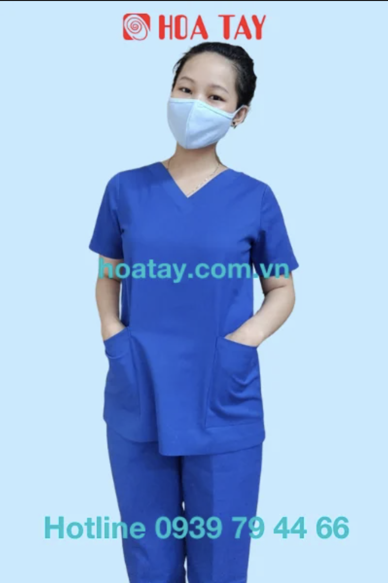 Một nữ nhân viên y tế đang đứng, mặc bộ đồ scrubs (đồng phục phẫu thuật) màu xanh dương, cổ chữ V, tay ngắn, có hai túi lớn phía trước, chất liệu vải trơn, phông nền xanh nhạt, ánh sáng studio đều.