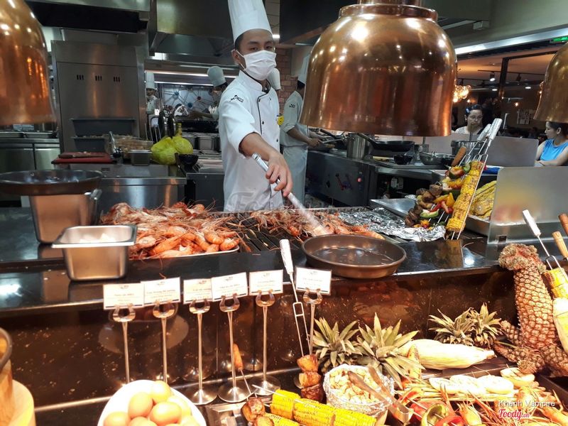 Một đầu bếp (nam) đang nướng tôm trên vỉ sắt; mặc áo khoác trắng cổ tàu (mandarin collar), đeo khẩu trang y tế, đội mũ toque cao; không gian bếp nhà hàng chuyên nghiệp với đèn sưởi đồng treo phía trên