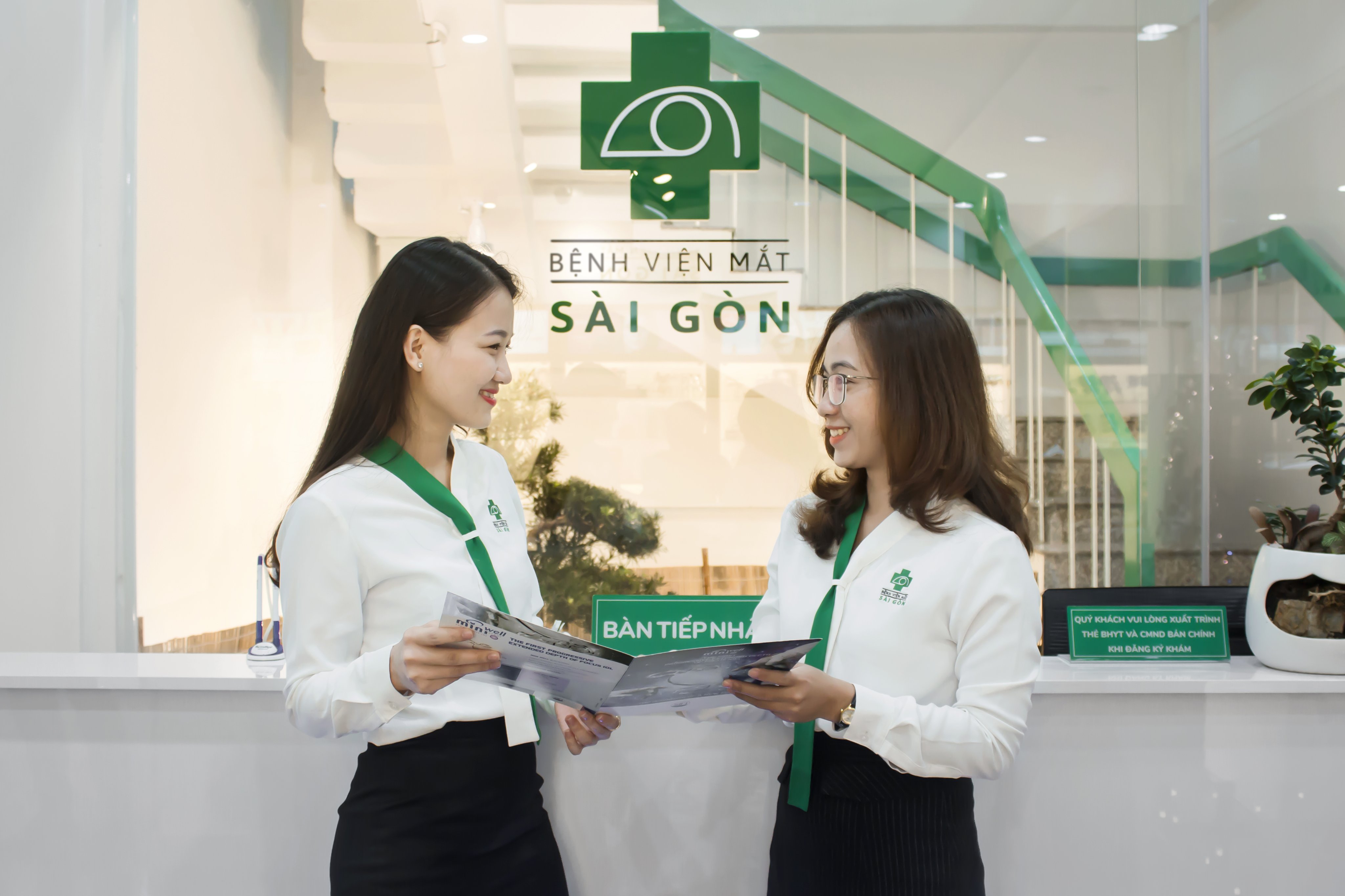 Hai nữ nhân viên -> trao đổi tài liệu -> áo sơ mi trắng cổ xanh lá, quần đen, logo bệnh viện -> quầy lễ tân kính, logo 'Bệnh viện Mắt Sài Gòn' phía sau, ánh sáng trắng