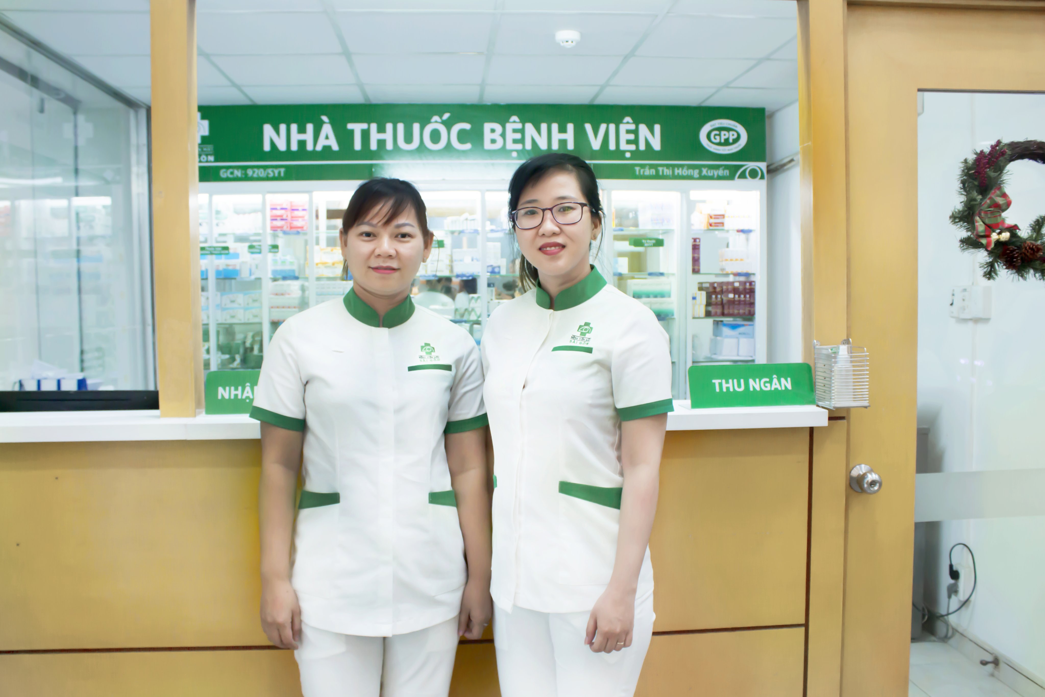 Hai nữ nhân viên đứng trước quầy thu ngân của nhà thuốc bệnh viện, mặc đồng phục trắng với cổ và tay áo viền xanh lá, logo thêu trên ngực, nền là tủ kính thuốc và biển hiệu xanh.
