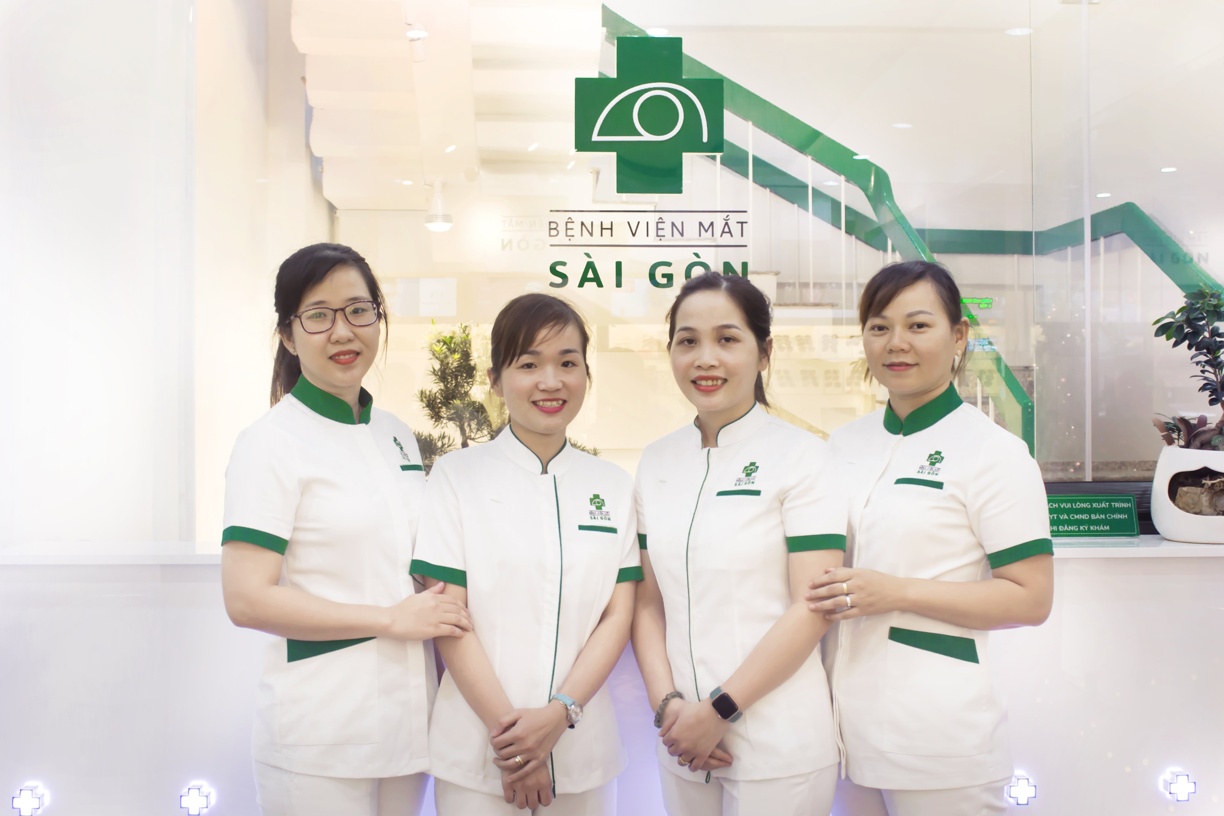 Bốn nữ nhân viên y tế mặc đồng phục trắng với viền cổ, tay áo và túi màu xanh lá cây, đứng trước quầy lễ tân có logo bệnh viện mắt Sài Gòn, ánh sáng trong nhà
